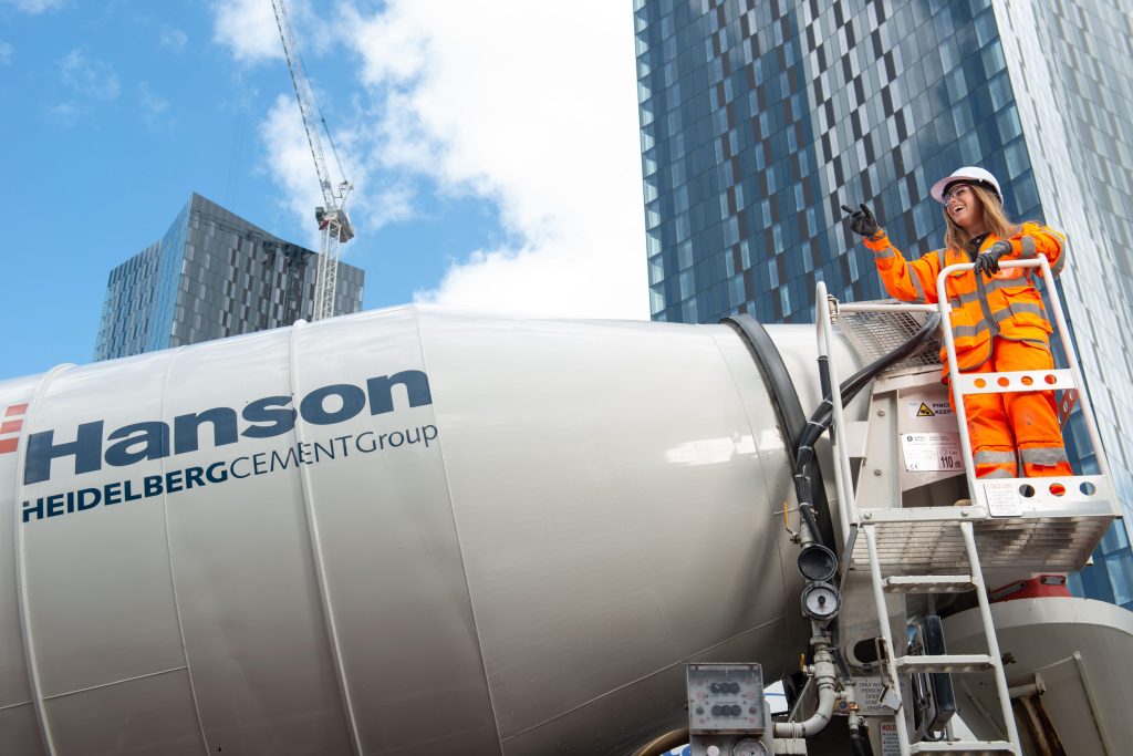 A woman in high-visibility orange workwear and a hard hat stands on a platform next to a Hanson cement truck, pointing forward, with tall modern buildings and a crane in the background.