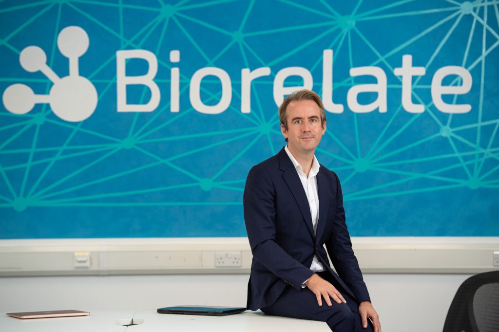 A man in a navy suit sits on a desk in front of a blue wall with the word Biorelate and a network graphic. A laptop and tablet are on the desk.