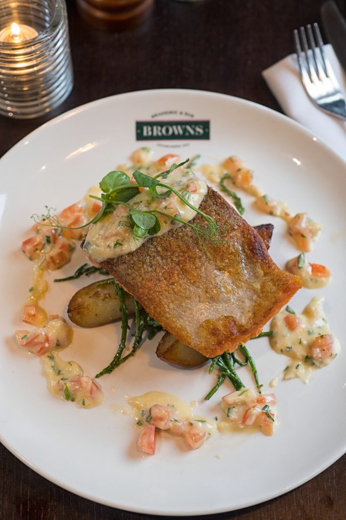 A plated dish featuring crispy-skinned fish atop roasted potatoes and green vegetables, garnished with a creamy sauce and diced tomatoes, served on a white plate labeled BROWNS. A lit candle and cutlery are nearby. Restaurant photography Manchester