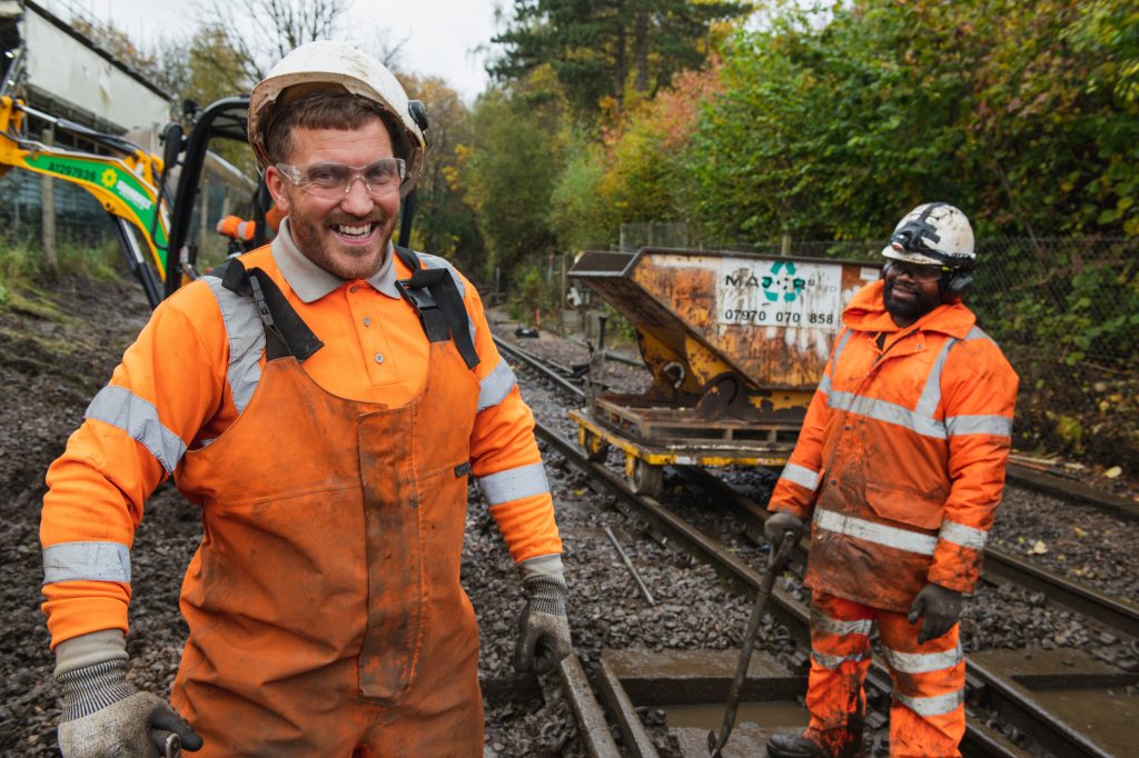 Two railway workers in orange safety gear and helmets stand on muddy train tracks; one smiles at the camera while the other holds a tool. There is construction equipment and autumn trees in the background. Industrial photography Greater Manchester -