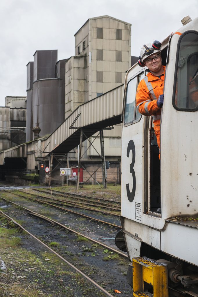 A worker in an orange safety suit and helmet leans out of a white train marked with the number 3. Industrial buildings and rail tracks are visible in the background under a cloudy sky. Engineering site photography Hope Valley