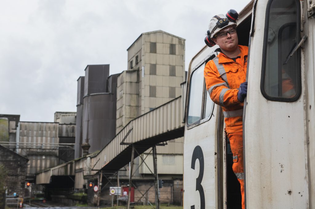 A worker in an orange jumpsuit and safety gear stands in the doorway of a white industrial train, numbered 3, at a large quarry & industrial facility with weathered buildings in the background. Engineering site photography Hope Valley