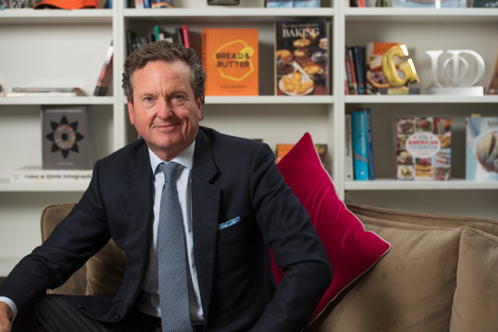 A man in a dark suit and tie sits on a brown couch with pink pillows, in front of a white bookshelf filled with books and decorative items. Previous CEO from St Pierre groupe ltd Headshot photographed at Didsbury, Manchester