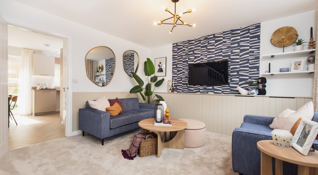 Modern living room with blue sofas, round mirrors, a geometric accent wall with a mounted TV, indoor plant, and decorative shelves. Natural light flows in from an open doorway leading to a bright kitchen area. Show house internal photography Greater Manchester