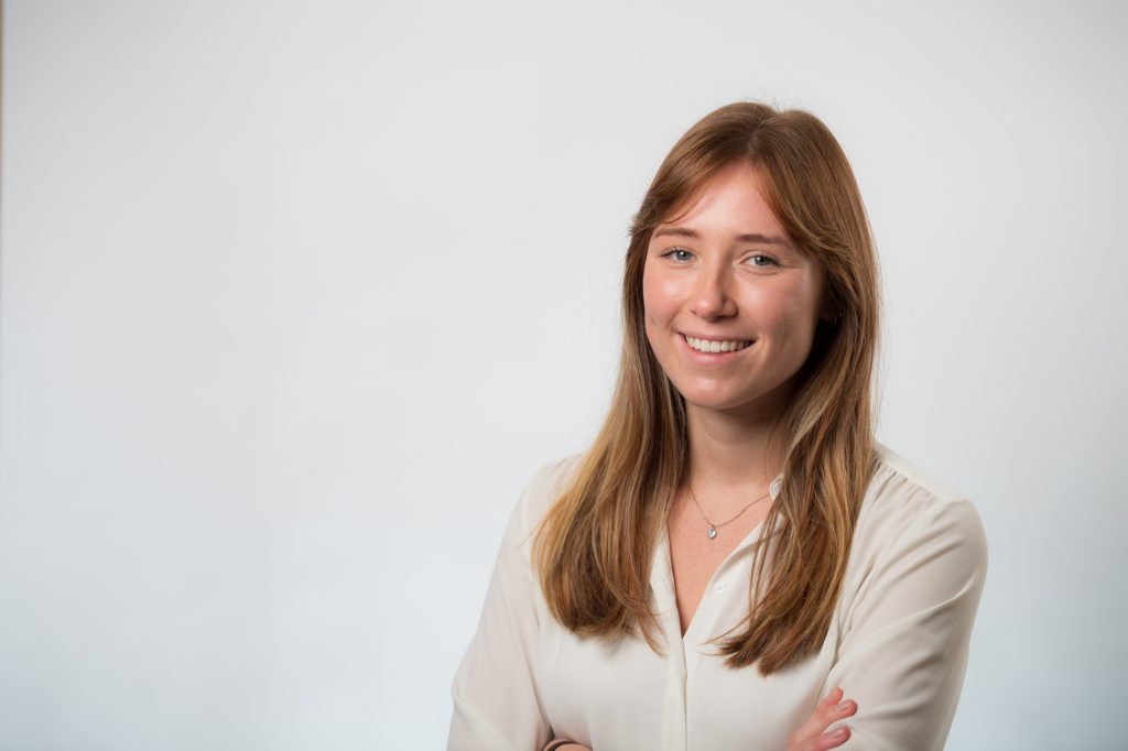 A young woman with long, light brown hair smiles at the camera, standing with arms crossed. She is wearing a white blouse and a necklace, with a plain white background. Headshot photographer Manchester
