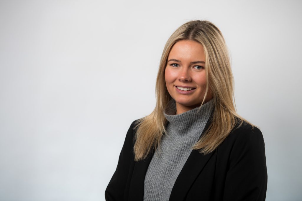A young woman with straight blonde hair, wearing a gray turtleneck sweater and a black blazer, smiles at the camera against a plain light background. Law firm headshot photography Sheffield