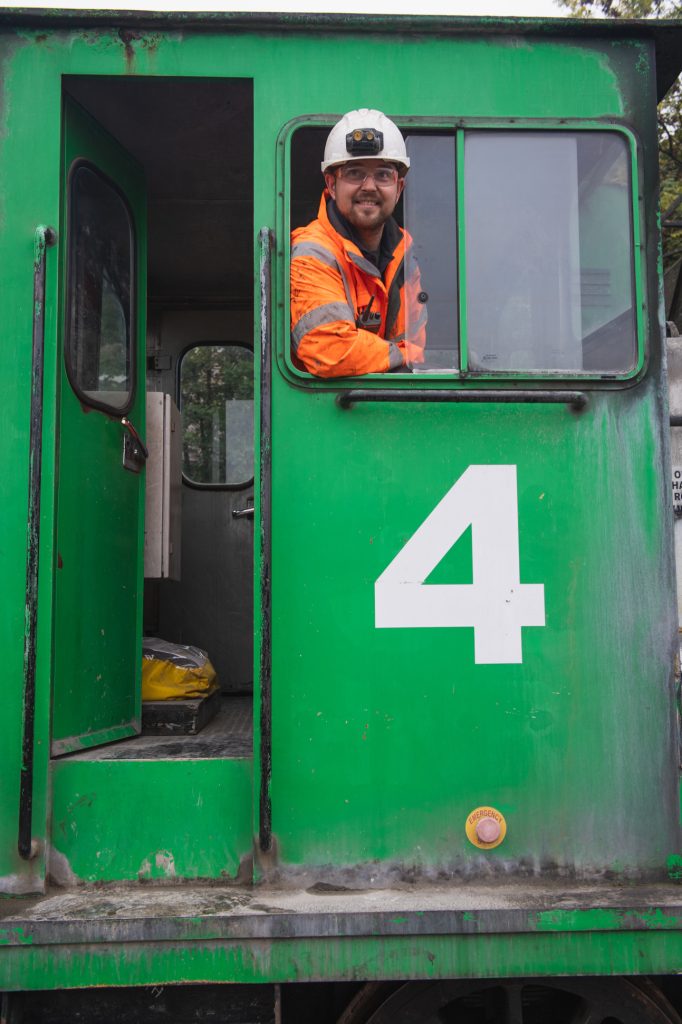 A man wearing an orange safety jacket and white hard hat smiles from the cab window of a green train engine marked with a large white number 4. Engineering firm photographer Sheffield