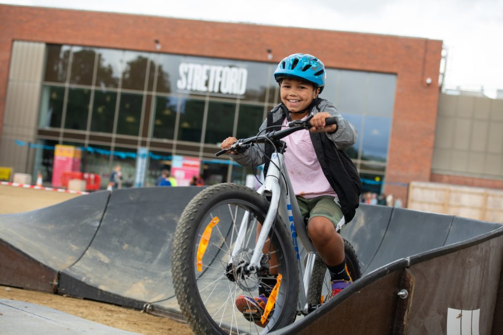 A young child wearing a blue helmet rides a bicycle over a curved ramp at an outdoor bike park, smiling. A modern building with large windows and the word STRETFORD is visible in the background. PR Photography at event Manchester