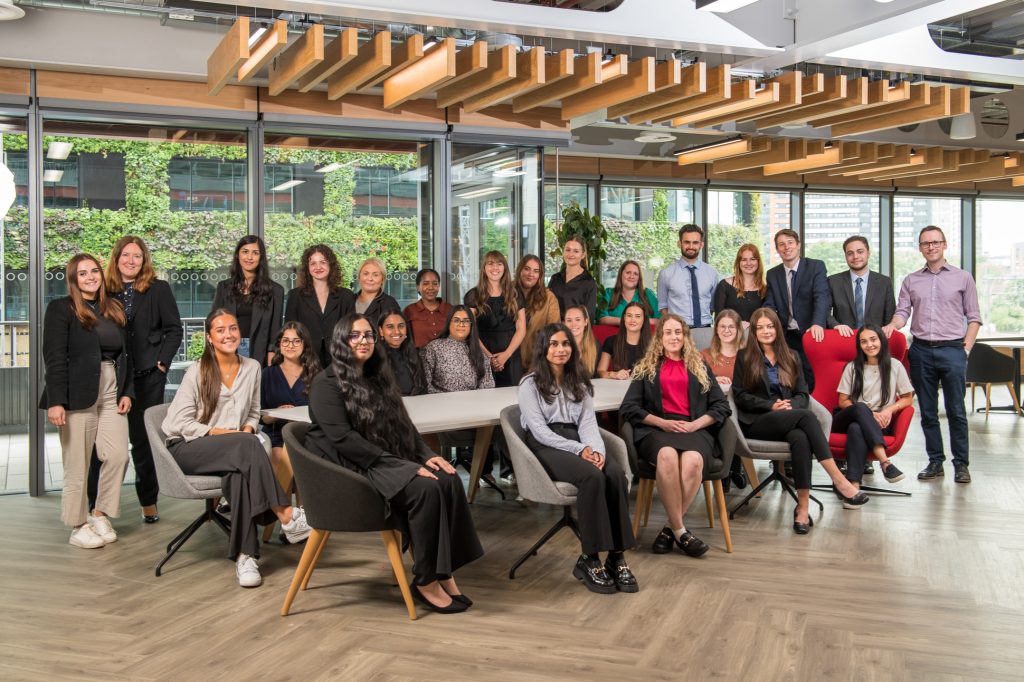 A group of around 25 people, dressed in business attire, pose for a photo in a modern office space with wooden floors, indoor plants, and large windows showing a green wall outside. Large team photography Manchester