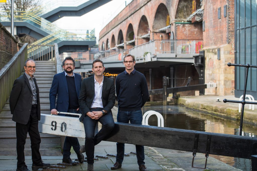 Four men in business attire pose by a canal lock in an urban area with brick arches, stairs, and water in the background. One man sits on the lock gate while the others stand nearby, all facing the camera. New team photography and headshots Manchester