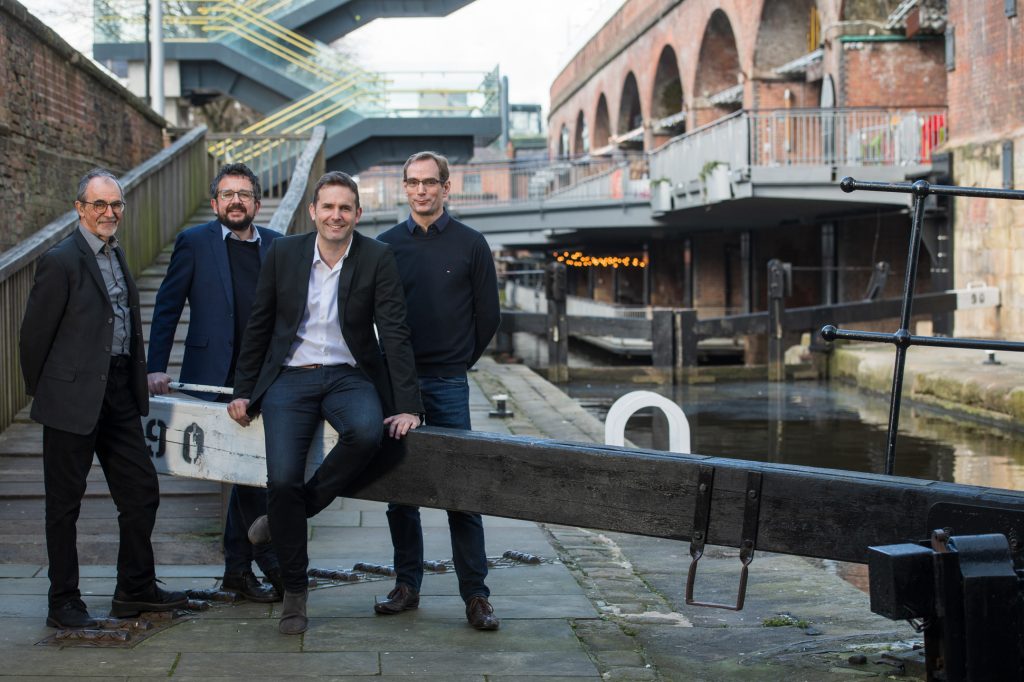 Four men in smart casual attire pose by a canal lock in an urban setting with brick buildings, metal railings, and staircases in the background. One man sits on a canal beam while the others stand nearby, smiling. New startup team photography and headshots Manchester