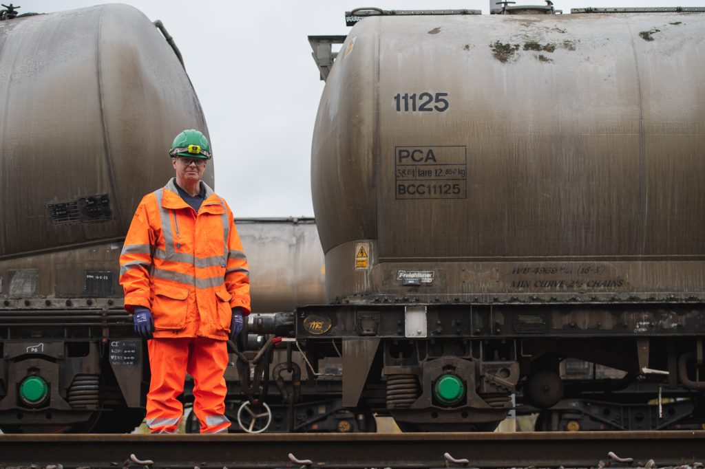 A worker in high-visibility orange safety gear and helmet stands on a railway track next to a large, weathered, cylindrical train tanker. The tanker is marked with codes and numbers. Industrial rail photography Hope Valley - Manchester Photographer