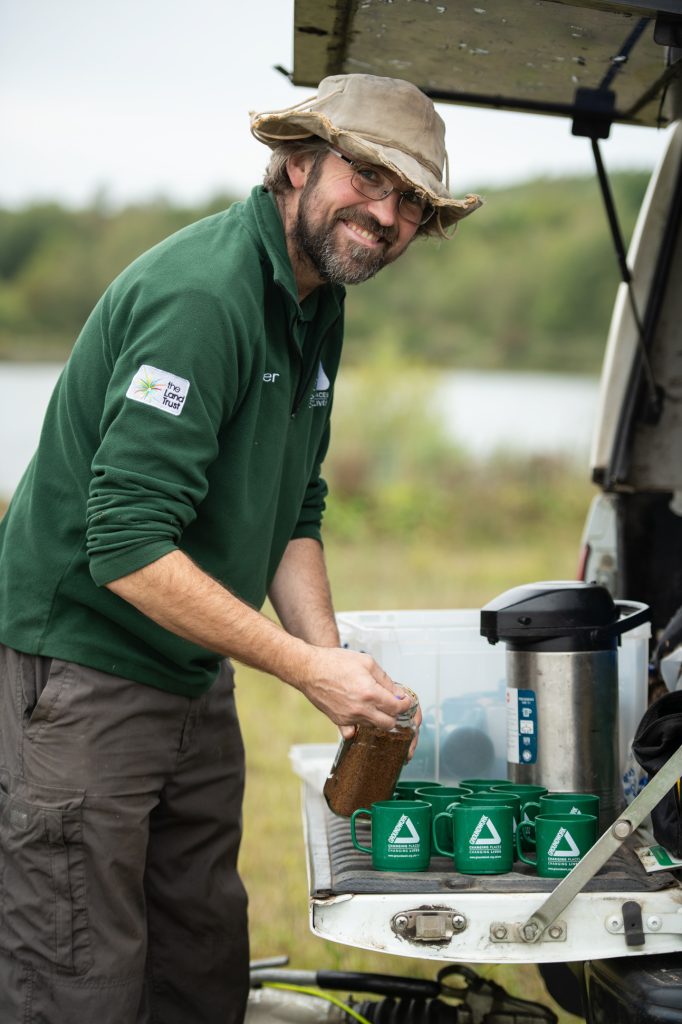 A man in a green jacket and wide-brim hat smiles while pouring coffee into green mugs from a thermos on the back of a vehicle in an outdoor setting near water and trees. the Land Trust event photography