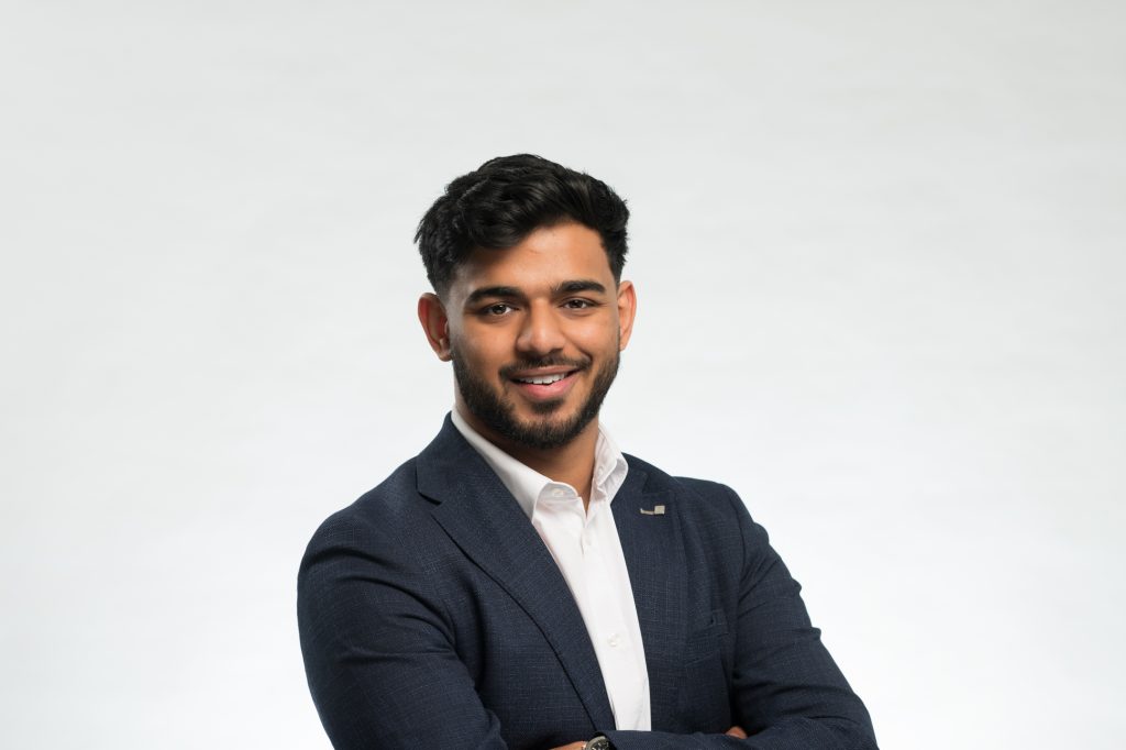 A man with short dark hair and a beard, wearing a navy blue suit and white shirt, stands smiling with his arms crossed against a plain white background. Headshot photography for Manchester law company plain background - Manchester Photographer