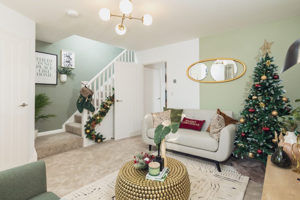 A modern living room decorated for Christmas with a tree, garland on the stairs, and stockings. Green accent wall, cream furniture, round gold coffee table, and wall art create a cozy, festive atmosphere. Show Home interior and new housing development photography in Lancashire by Manchester Photographer