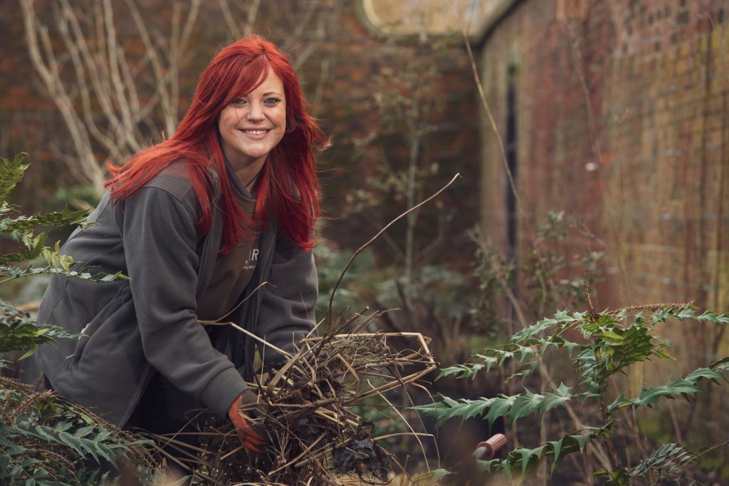 A woman with long red hair smiles while gathering dry branches and twigs in a garden area with overgrown plants and brick walls. She is wearing a grey jacket and gloves. RHS Bridgewater Garden Manchester PR photography