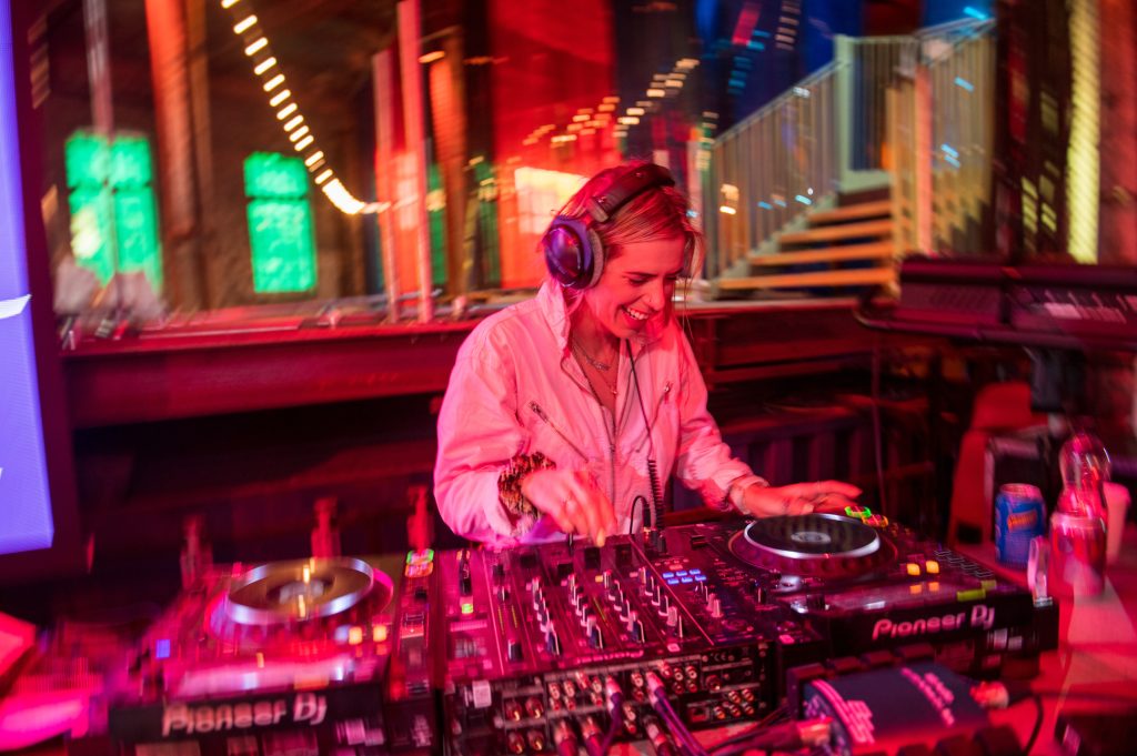 A smiling DJ wearing headphones mixes music on a Pioneer DJ controller at a vibrant, colorful venue with blurred lights and reflections in the background. Corporate event photography at Liverpool