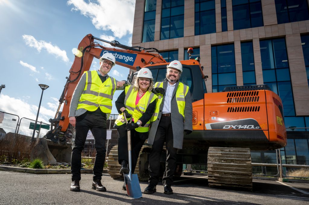 Three people wearing high-visibility vests and hard hats smile and pose in front of an orange excavator and a modern office building; one holds a shovel, groundbreaking event photography Manchester
