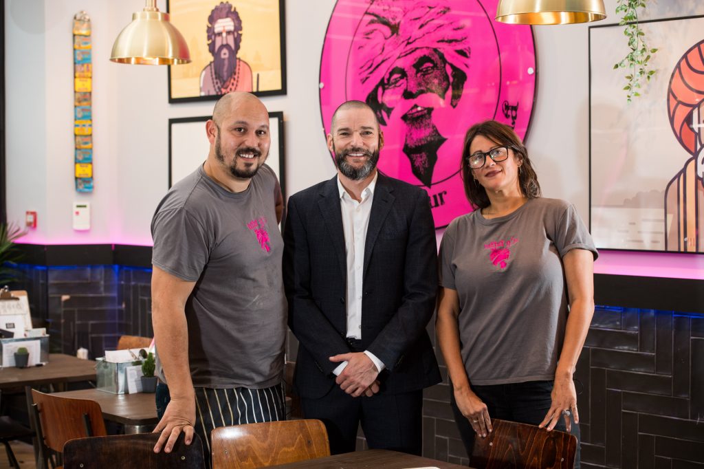 Three people pose in a restaurant: two staff in matching gray t-shirts with a pink logo, standing on either side of a man in a dark suit. Colorful, Indian-themed artwork decorates the walls behind them. Million pound menu restaurant with Fred Sirieix- PR photography Manchester