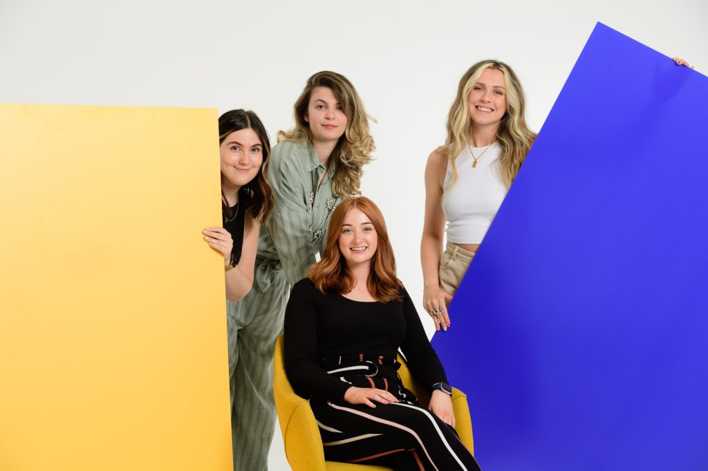 Four women pose together in a bright studio. One sits on a yellow chair, while three stand behind large yellow and blue panels. All are smiling and dressed in casual, stylish clothing against a white background. Manchester photographer cost to book professional photography