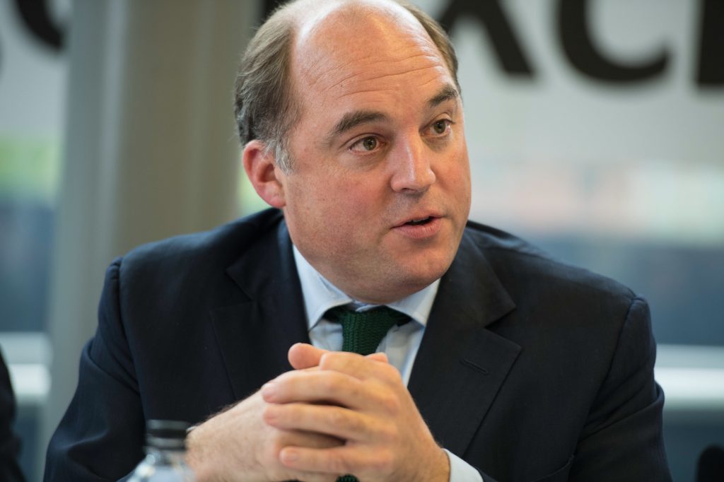 A man in a suit and tie sits at a table, clasping his hands and speaking. The background is blurred with large letters partially visible.