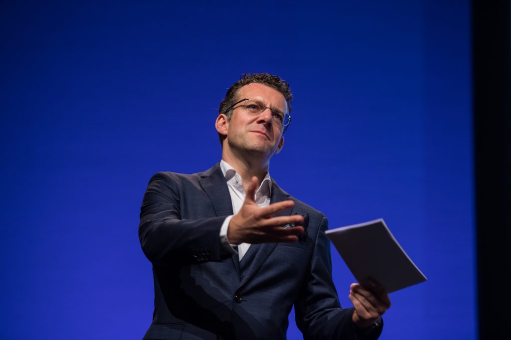 A man in a dark suit and glasses gestures with one hand while holding papers in the other, standing in front of a plain blue background, appearing to speak or present. Conference photography and video Mancheser