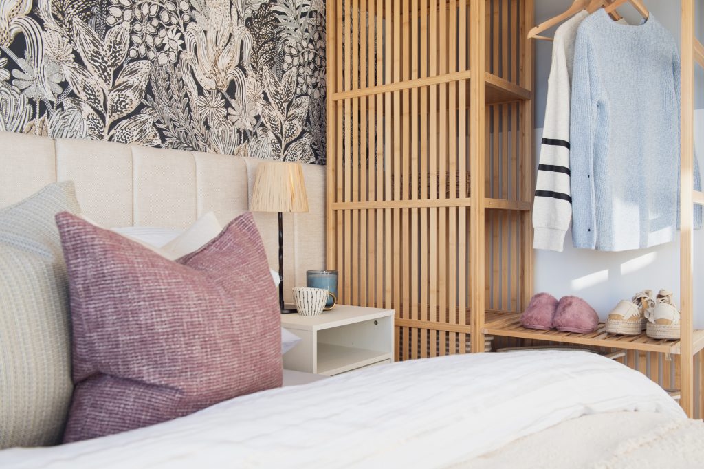 A cozy bedroom features a bed with pastel pillows and white bedding, a wooden nightstand with a lamp and mug, a wooden slatted wardrobe, clothes on hangers, and shoes neatly arranged. Botanical wallpaper decorates the wall. Hive Homes new housing development - show home photography Manchester
