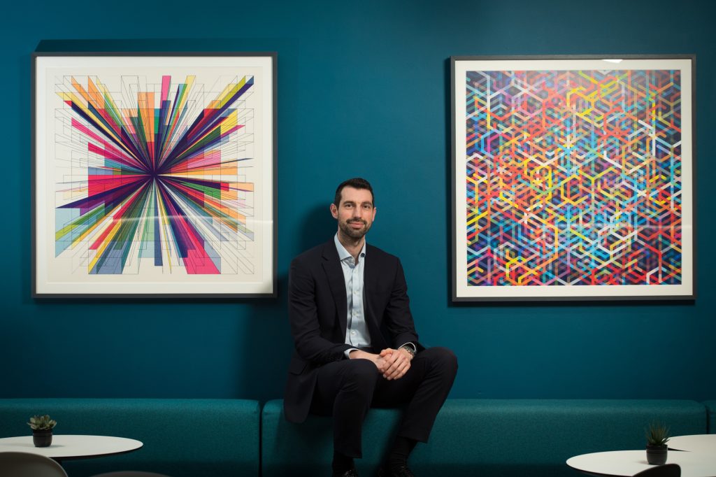 A man in a dark suit sits on a teal bench between two colorful abstract artworks, against a teal wall. Small potted plants and tables are in the foreground. Corporate business portrait Manchester
