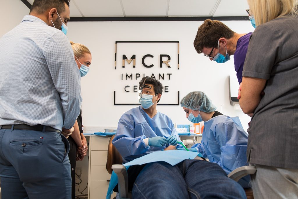 A group of dental professionals wearing masks and scrubs gather around a patient seated in a dental chair at MCR Implant Centre, observing a procedure in progress. Case study photography Manchester