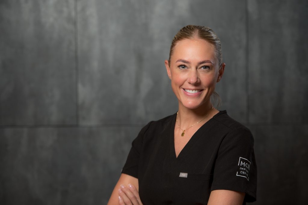 A woman in black medical scrubs stands smiling with arms crossed in front of a gray, textured background. She has blonde hair pulled back and wears a gold necklace. The sleeve has a white logo and text. Manchester Implant Centre headshot photography