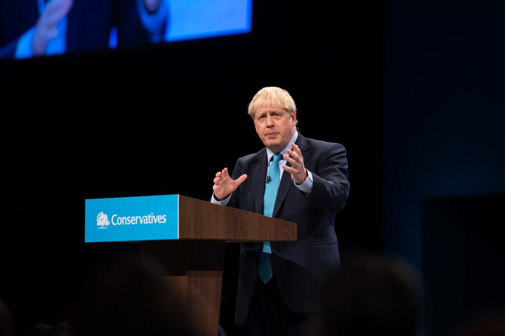 A man in a suit and blue tie stands at a podium labeled “Conservatives,” speaking and gesturing with his hands during a formal event or conference. Boris Johnson speaking at Tory party conference - conference photography Manchester Central exhibition centre.