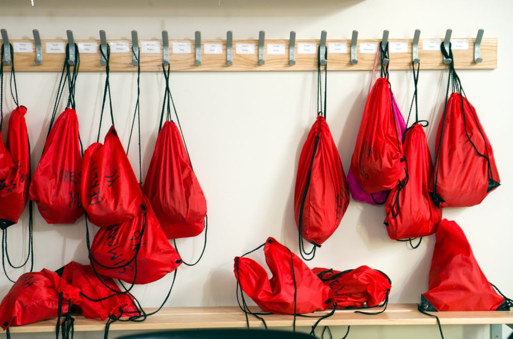 Several red drawstring bags hang on hooks on a wall-mounted rack, with some bags resting on a wooden shelf below. Each hook has a small label above it. One pink drawstring bag is mixed among the red ones. New school builder photography for annual report