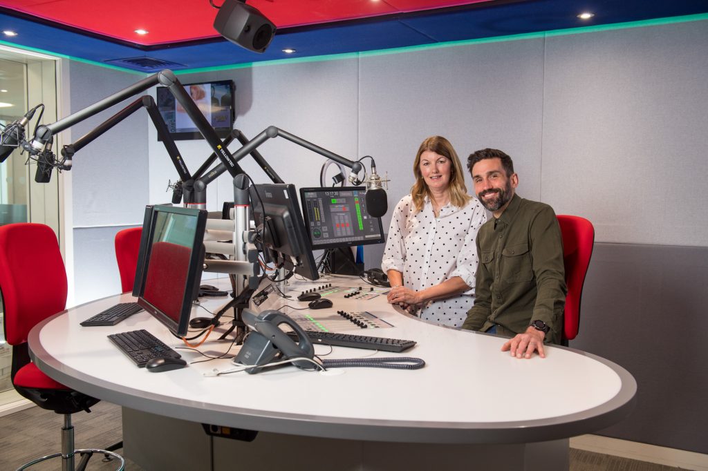 Two people, a woman and a man, sit together in a modern radio studio with microphones, computer monitors, and red chairs. The woman stands while the man sits, both smiling at the camera. Podcast photography Manchester