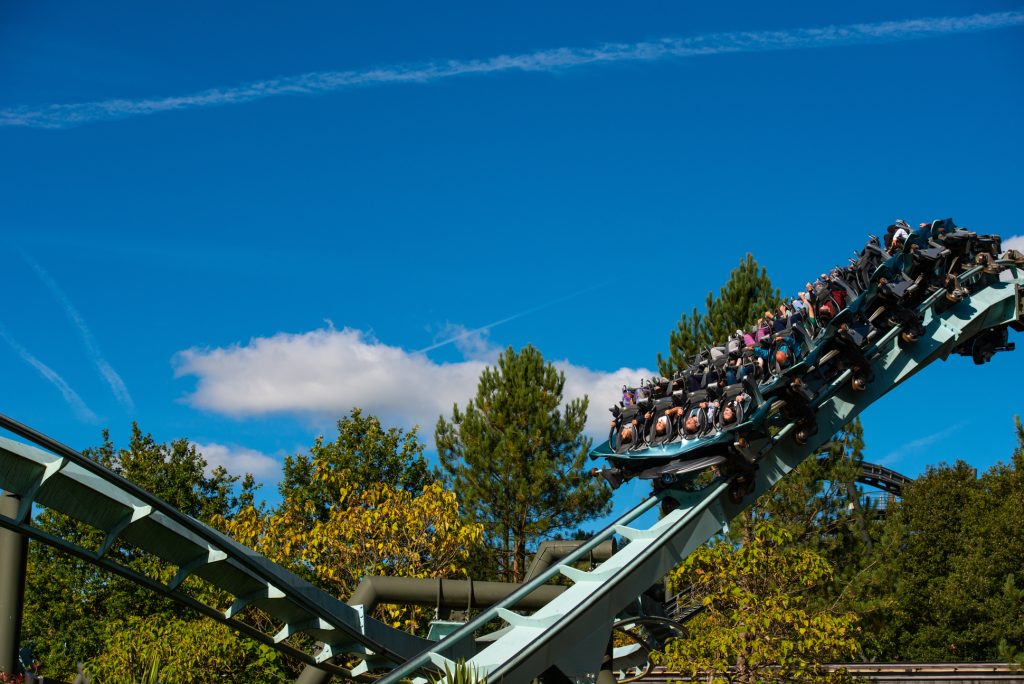 People ride a looping roller coaster on a sunny day, surrounded by green trees and a bright blue sky with a few white clouds and contrails in the background. Alton Towers marketing photography Staffordshire