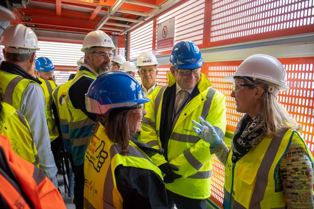 A group of people wearing hard hats, safety vests, and protective gear stand and talk inside a construction site, surrounded by red metal mesh walls and sunlight filtering through. Property developers with shareholders PR photography Leeds