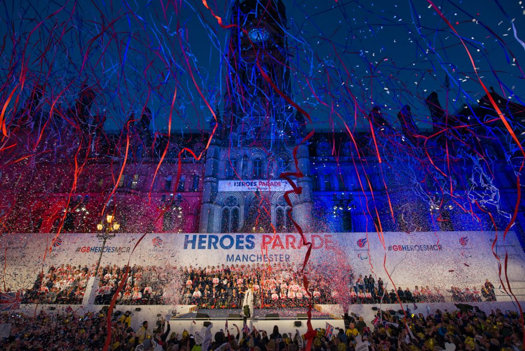 A large crowd celebrates at an outdoor Heroes Parade in Manchester, with red, white, and blue confetti and streamers filling the air in front of a decorated historic building at night. Event photography for press Manchester