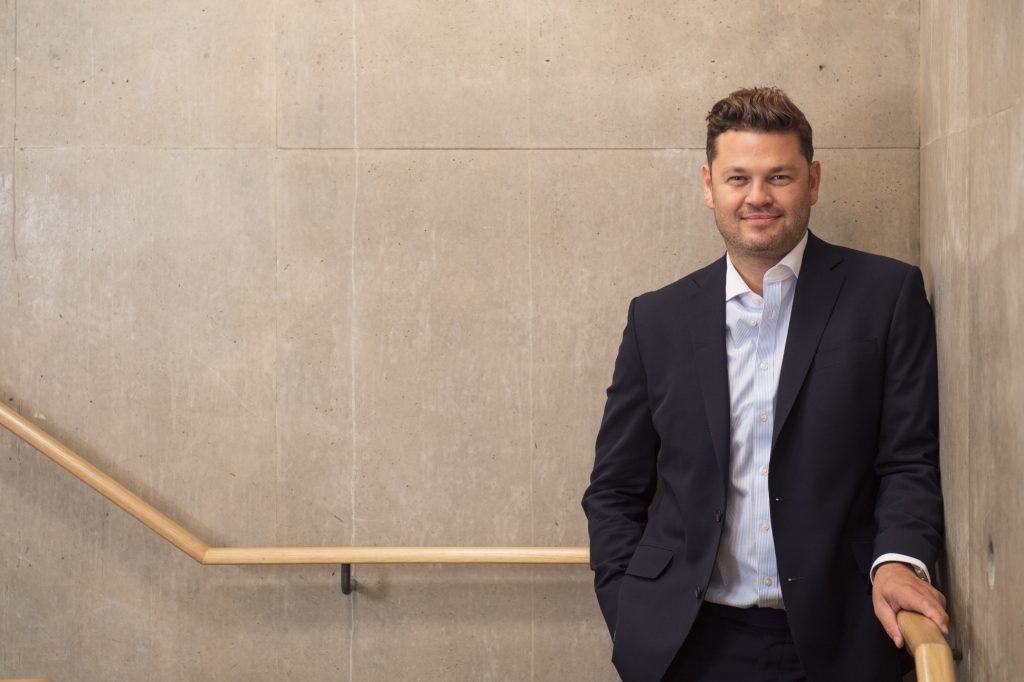 A man in a dark suit stands by a light wooden handrail against a plain, light-colored concrete wall, smiling with one hand in his pocket. relaxed headshot for law firm regional offices