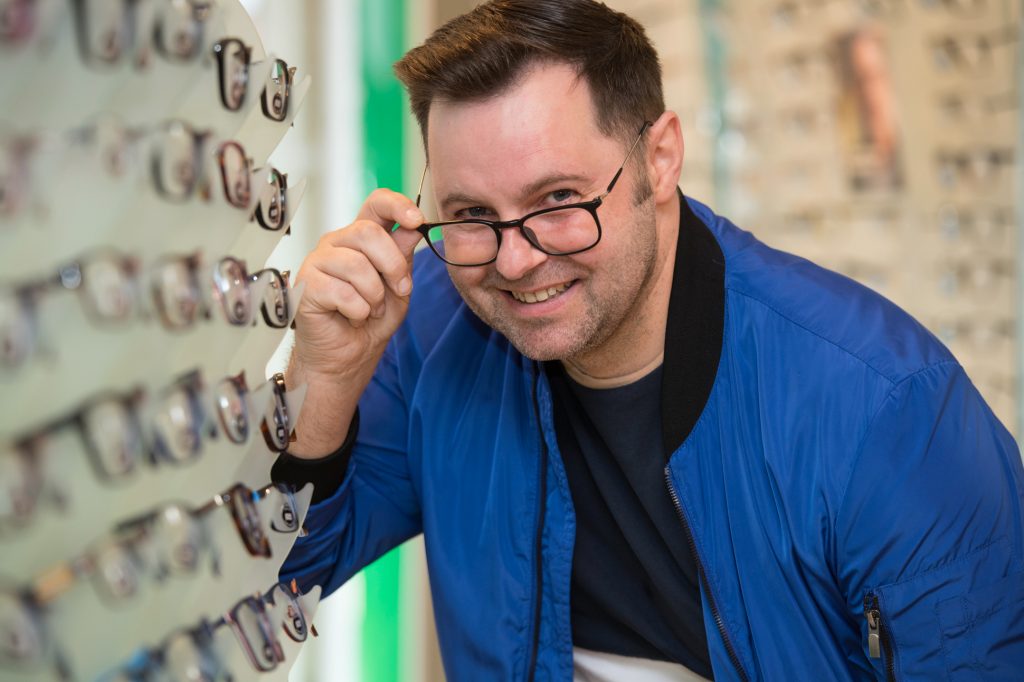 A smiling man in a blue jacket tries on glasses in an eyewear store, lifting the frames slightly while standing next to a display of various eyeglasses. PR photography Specsavers at Bolton