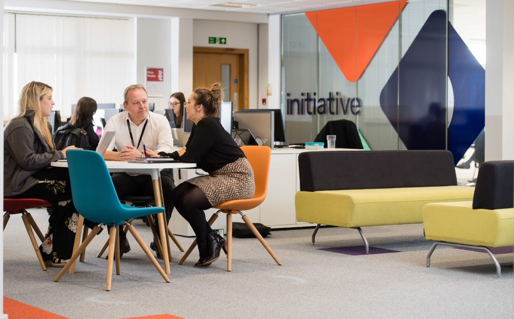 Four people sit around a table in a modern office, engaged in conversation. Colorful chairs and a yellow sofa are visible, with a glass wall behind them displaying the word initiative. Corporate case study photographer UK