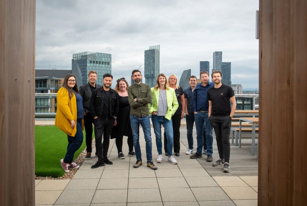 A group of ten people stands on a rooftop terrace with a city skyline in the background. They are smiling and dressed in casual clothes, framed by two wooden structures on either side. Relaxed team photography and headshots Manchester