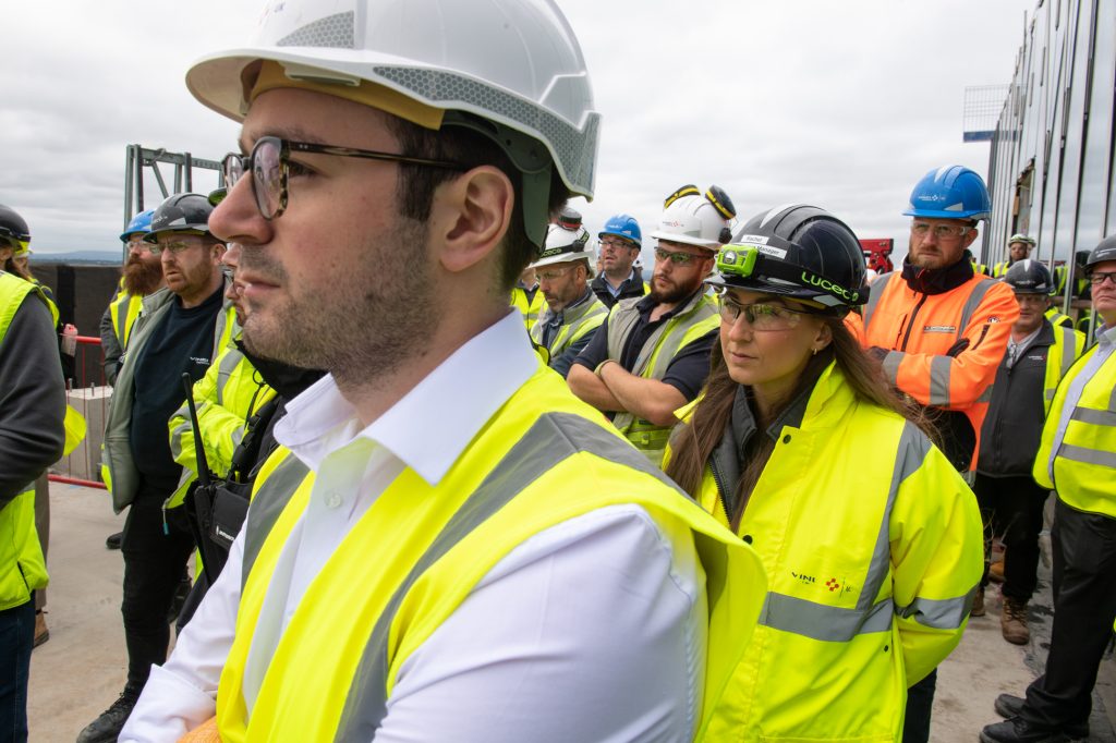 A group of construction workers wearing hard hats, safety glasses, and high-visibility jackets stand outdoors, listening attentively. The sky is overcast and the background shows construction materials and structures. Construction site photographer Greater Manchester
