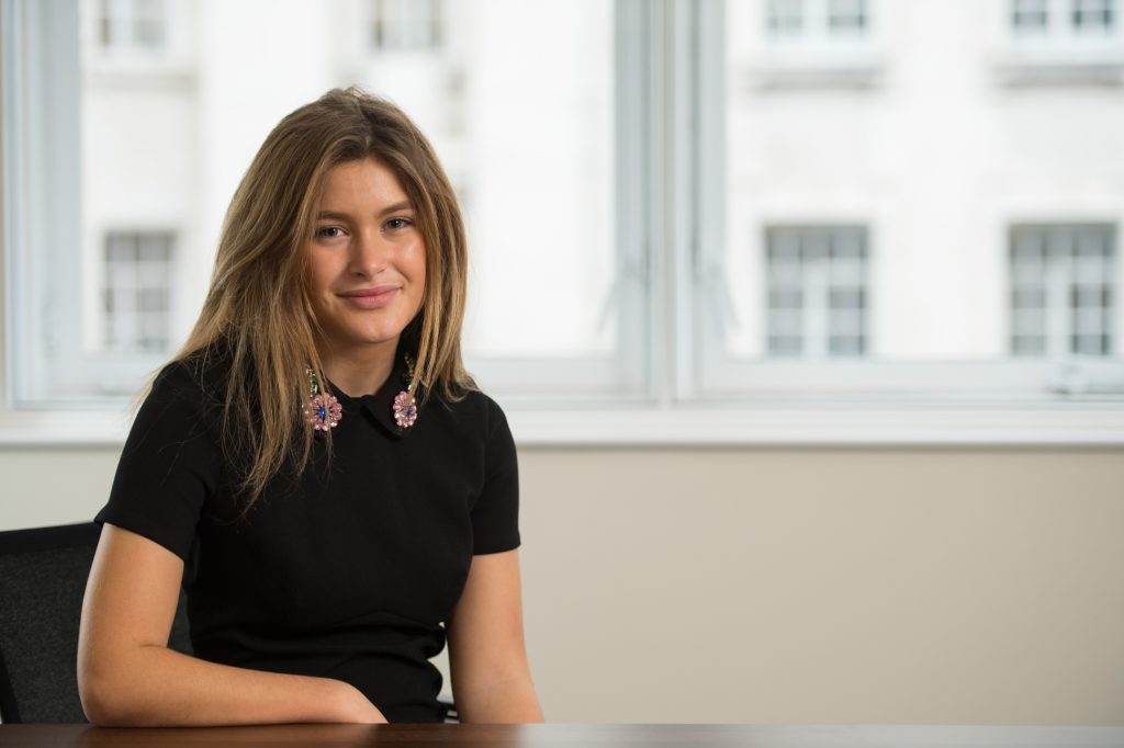 A young woman with long brown hair, wearing a black top and large decorative earrings, sits at a wooden desk in a bright office with large windows in the background. Headshot photographer Manchester