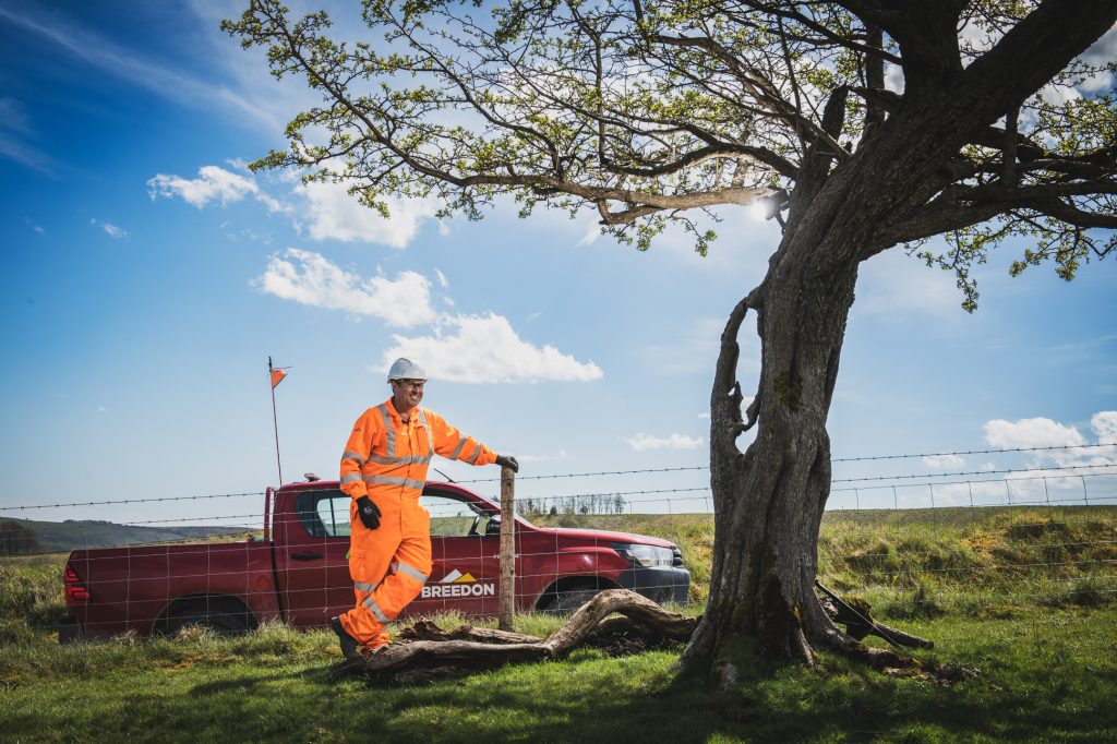A worker in an orange high-visibility suit and white helmet stands by a tree and a fence, next to a red pickup truck, under a blue sky with scattered clouds. Industrial photographer Derbyshire