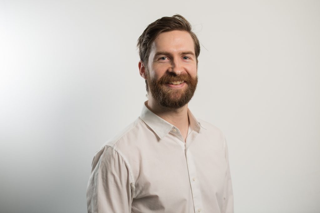 A man with a beard and mustache, wearing a white button-up shirt, smiles while standing against a plain white background. Headshots manchester