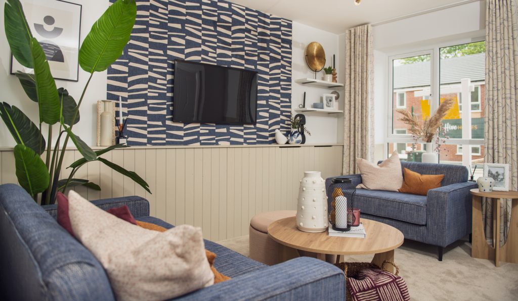 A modern living room with blue sofas, patterned wallpaper, a wall-mounted TV, houseplants, and a round wooden coffee table with decorative items, near a large window with beige curtains letting in natural light. New housing development photography