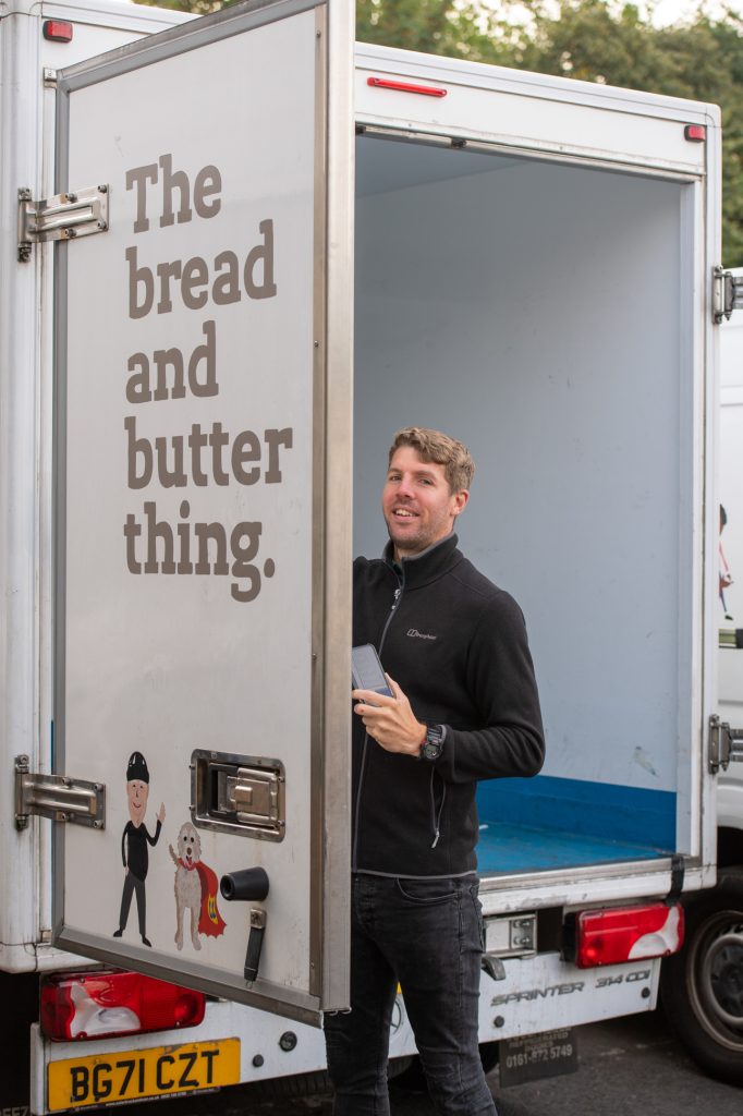A man stands smiling by the open door of a white delivery van with The bread and butter thing written on the side, holding a phone. The van is mostly empty and the license plate reads BG71 CZT. Charity PR photography Greater Manchester