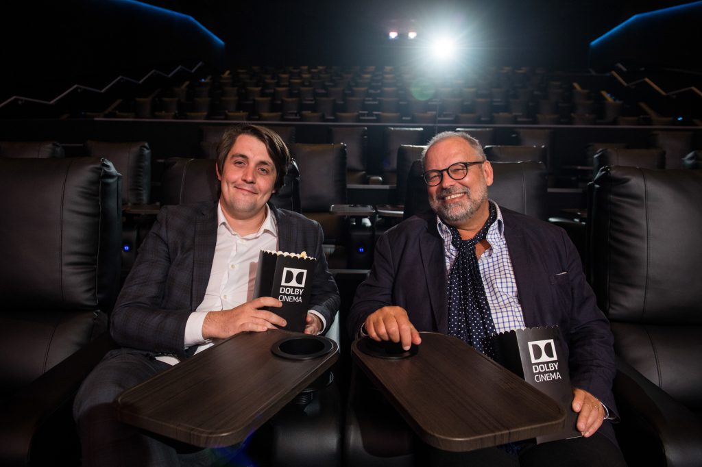 Two men sit in reclining seats at a Dolby Cinema, each holding a box of popcorn, smiling at the camera with empty theater seats and bright lights visible behind them. DCM event photography Leeds