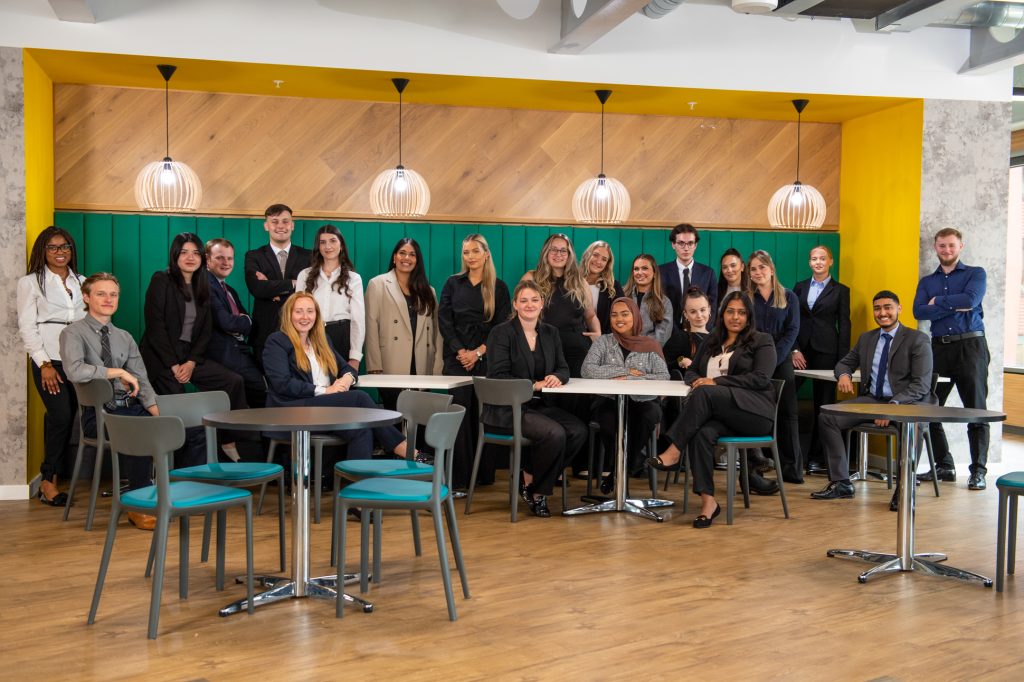 A group of professionally dressed people pose and smile in a modern office break area with wooden floors, green seating, and yellow walls. Some are standing behind, while others are sitting at tables in front. Corporate team photography Greater Manchester