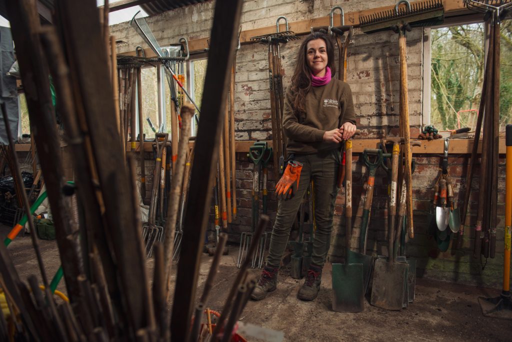 A woman in work clothes stands in a shed filled with various gardening tools hanging on the wall. She is smiling, holding gloves, and surrounded by rakes, shovels, and spades. Sunlight shines in through the windows behind her. RHS Manchester event photography