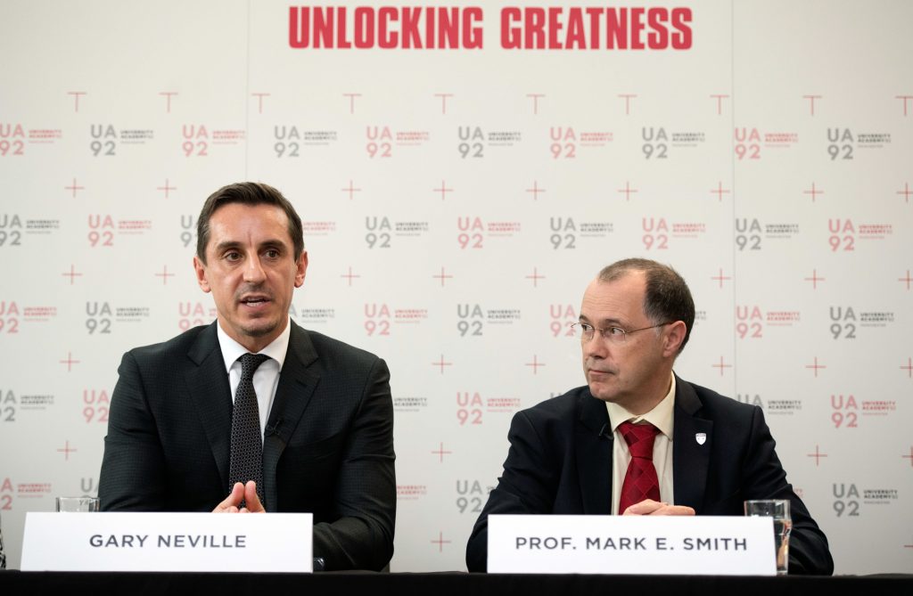 Two men in suits sit at a table with name plates reading Gary Neville and Prof. Mark E. Smith in front of a backdrop displaying UA92 and Unlocking Greatness. Gary Neville is speaking; Mark E. Smith looks on. Event photography Manchester